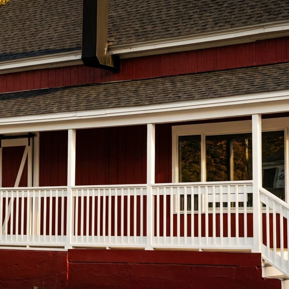 Red building with a white porch, railing, and steps, featuring a dark roof and windows.