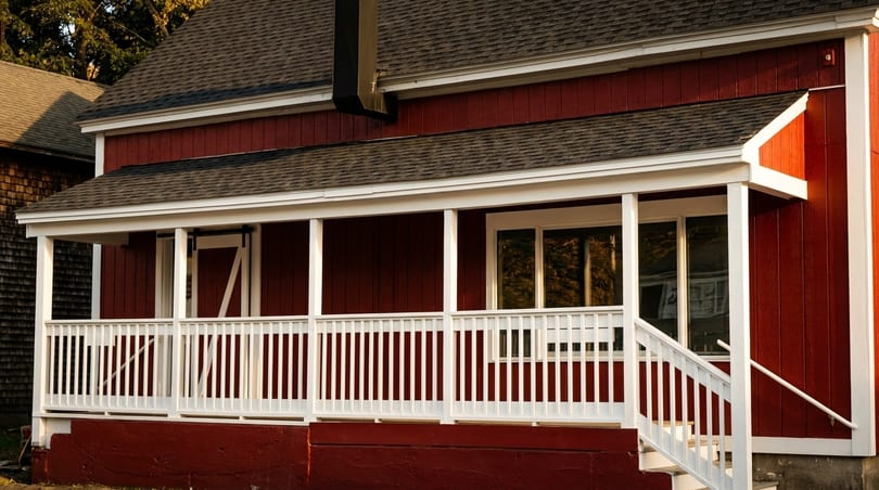 Red building with a white porch, railing, and steps, featuring a dark roof and windows.