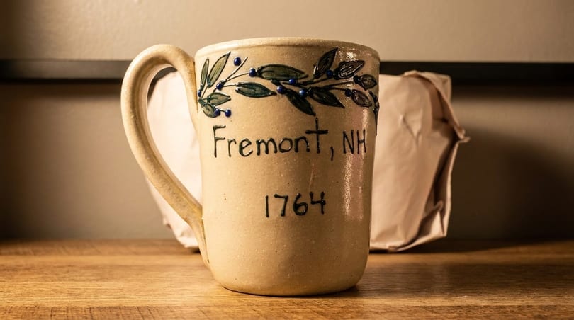 A ceramic mug with "Fremont, NH 1764" and a leaf design, on a wooden surface.